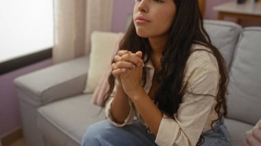 Uzun esmer saçlı genç bir kadın yumuşak ışıklandırması ve rahat bir atmosferi olan rahat bir oturma odasındaki kanepede düşünceli bir şekilde oturuyor.