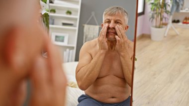 Üstsüz, gri saçlı yaşlı bir adam modern yatak odasının aynasında cilt bakımı uyguluyor..