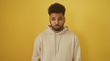 Gayri resmi bej kapüşonlu yakışıklı Afro-Amerikan bir adam, parlak sarı bir arka plana karşı poz veriyor, düşünceli görünüyor..