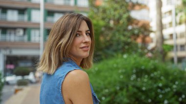 Şehir parkında gülümseyen güzel sarışın kadın, şehirde sakin bir an sergiliyor. Arka planda binalar ve yeşillikler var..