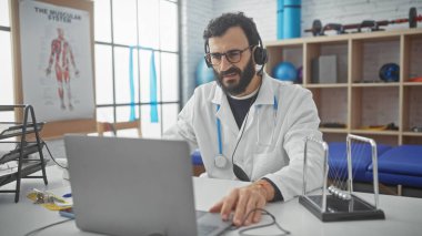 Laboratuvar önlüklü, orta yaşlı sakallı bir adam hastane ofisinde dizüstü bilgisayarla çalışıyor. Masasında Newton beşiği var..