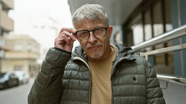 Kendine güveni tam orta yaşlı, gri saçlı ve gözlüklü bir adam şehir sokağında dikiliyor, gözlerini düzeltiyor, doğrudan kameraya bakıyor.