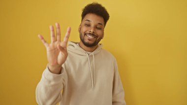 Kapüşonlu neşeli yetişkin bir siyah adam, sarı izole edilmiş arka planda parmaklarıyla dört numarayı gösteriyor..