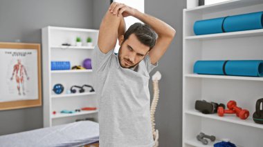 Handsome hispanic man stretching in a modern rehabilitation clinic's gym, indicating a focus on physical therapy and wellness. clipart