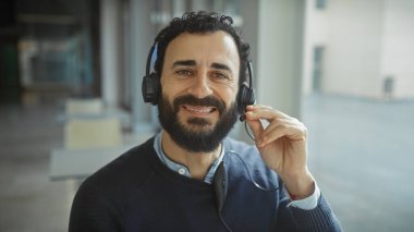 Modern ofiste kulaklıklı, gülümseyen sakallı bir adam, günlük profesyonellik ve yaklaşabilirlik yayıyor..