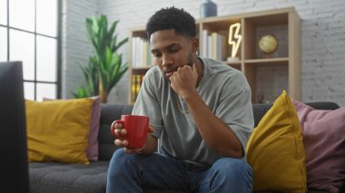 Thoughtful african american man holding mug in modern living room. clipart