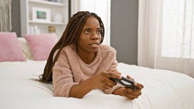 Kafasında bornoz olan odaklanmış siyah bir kadın rahat bir yatak odasında video oyunu oynuyor..