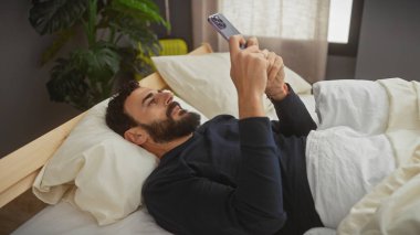 Sakallı adam, modern bir yatak odası ortamında akıllı telefon kullanarak yatakta yatıyor..
