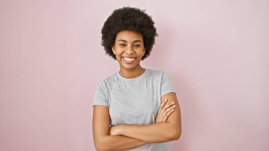 Kıvırcık saçlı ve kollarını kavuşturmuş gülümseyen Afro-Amerikan kadın pembe bir duvara yaslanmış, özgüvenini resmediyor..
