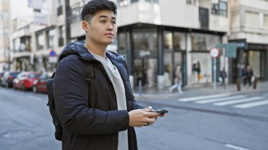 Akıllı telefonlu genç Asyalı bir adam bir şehir caddesinde duruyor, şehir yaşam tarzını canlandırıyor..