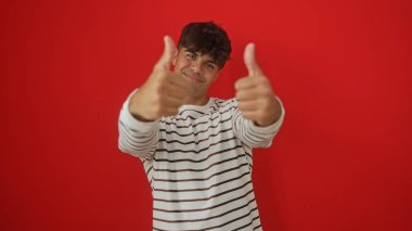 Yakışıklı, genç, İspanyol asıllı, kendine güvenen, çizgili gömlek giyen ve canlı kırmızı bir arka planın önünde duran bir adam..