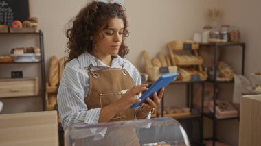 Fırında çalışan genç bir kadın elinde tabletle ekmek ve hamur işleriyle dolu rafların arasında duruyor.