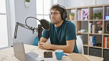 Sakallı genç bir İspanyol adam, iyi aydınlatılmış bir radyo stüdyosunda bir podcast sunuyor, dizüstü bilgisayar ve mikrofon ile donatılmış..