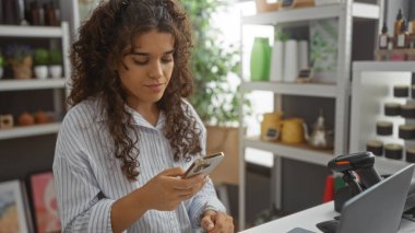 Esmer saçlı genç bir kadın modern iç mimarisi ve çeşitli dekorasyonları olan bir mağazada akıllı telefon kullanıyor.
