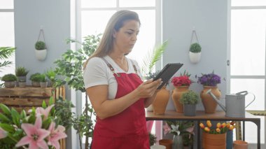 Kırmızı önlüklü, odaklanmış bir İspanyol kadın, kapalı alandaki bitkilerin arasında bir tablet kullanıyor. Huzurlu bir ev bahçesi atmosferi yaratıyor..