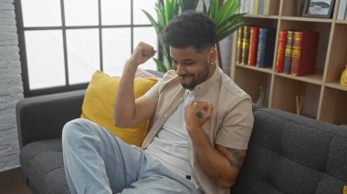 Gülümseyen Afrikalı Amerikalı adam modern bir oturma odasında kitap ve bitkilerle dinleniyor..