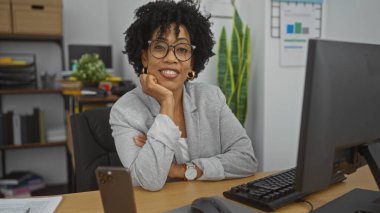 Kıvırcık saçlı, ofis ortamında güvenle gülümseyen, bilgisayarlı bir masada çalışan, gözlük takan ve iş kıyafeti giyen Afro-Amerikan genç bir kadın..
