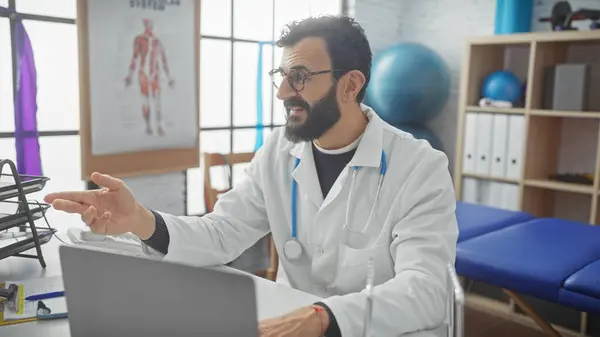 stock image Bearded doctor in glasses gesturing during a consultation in a clinic's examination room with a laptop.