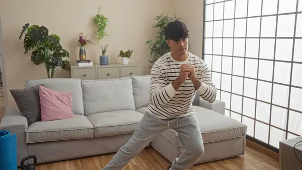 stock image A handsome young hispanic man exercising indoors in the living room of his modern apartment, with plants and contemporary furniture in the background.