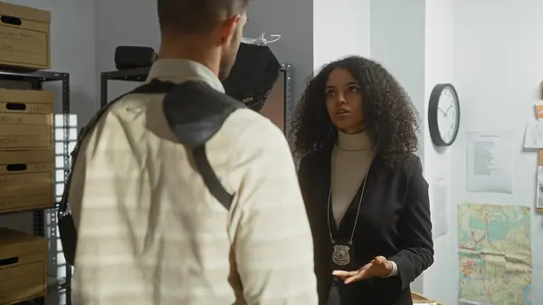 A woman detective with a badge converses with a man in a cluttered police department office, filled with evidence and documents.