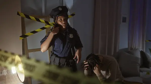 stock image A policewoman talks on a walkie-talkie at a night-time indoor crime scene with caution tape as a distressed man sits nearby.