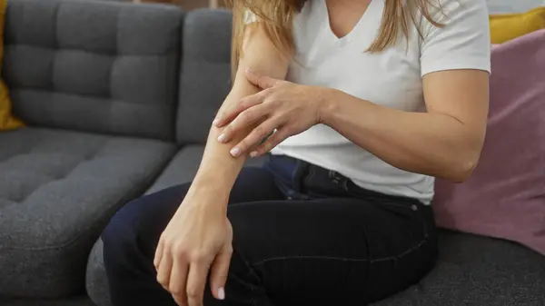 stock image A young woman sitting indoors scratches her arm while relaxing on a modern gray couch, showcasing everyday home life.