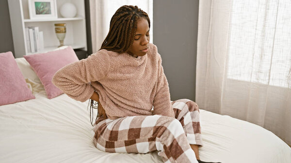 Красивая африканская американка с косичками сидит с болью на кровати в помещении, демонстрируя концепцию здоровья в домашней обстановке.