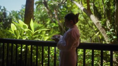 Bali Resort 'un yemyeşil yeşiliyle çevrili terasta kahve içen genç bir kadın, sakin tropikal bir sabahı kucaklıyor..
