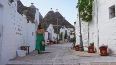 Yeşil elbiseli genç bir İspanyol kadın İtalya, Alberobello 'nun tarihi caddelerinde geziniyor, büyüleyici trulli evleri ve canlı saksı çiçekleri ile çevrili..
