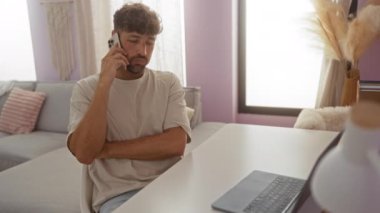 Oturma odasındaki rahat bir odada akıllı telefondan konuşan genç bir adam. Masasında dizüstü bilgisayarı ve sıradan dekore edilmiş bir apartman dairesi var.