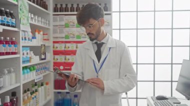 Sakallı, gözlüklü, beyaz laboratuvar önlüğü ve kravat takmış genç, İspanyol bir adam, ürünlerle dolu bir eczanede bir panoyu inceliyor..