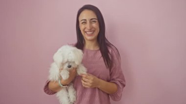 Genç, kendinden emin ve mutlu, dişlek, çekici bir gülümsemesi olan, pembe bir arka planda duran, neşeli bir şekilde sevgili köpeğine bakan İspanyol bir kadın.