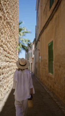 Fornalutx, Mallorca 'da güneşli bir sokağı keşfeden genç bir kadın, geleneksel Akdeniz mimarisi ve bereketli palmiye ağaçları ile çevrili, yazlık giysiler ve şapka giyiyor.