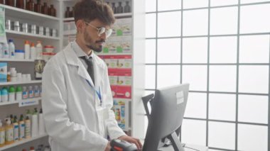 Sakallı, gözlüklü ve beyaz önlüklü genç bir İspanyol, parlak bir eczanenin içindeki bir bilgisayarda çalışıyor.