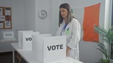 Genç, çekici bir İspanyol kadın İrlandalı bir seçim odasında, profesyonel giyinmiş, oy verme kabinlerinin yanında gülümserken başparmağını kaldırıyor..