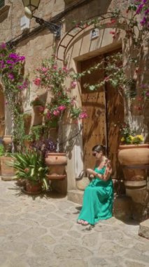 Yeşil elbiseli genç bir kadın Valldemossa, Mallorca 'daki kaldırım taşı caddesinde sepetle oturuyor, berrak güneşli gökyüzünün altında, kırsal bir binanın yakınındaki canlı saksılarla çevrili.