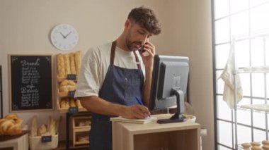 Pastanedeki genç adam önlük takıyor ve önünde bilgisayar ekranı olan bir tezgahta notlar yazarken telefonla konuşuyor.