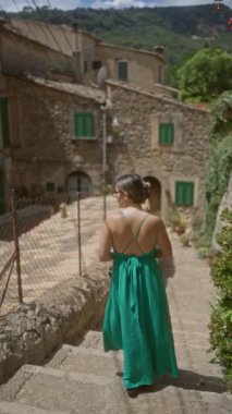 Yeşil elbiseli bir kadın Valldemossa, Mallorca 'da taş basamaklardan iniyor. Güneşli bir günde. Arka planda geleneksel evler ve yeşil pencere panjurları var..