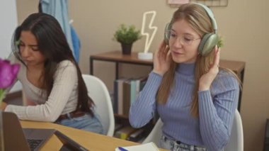 İki kadın ev ortamında, biri kulaklıkla dans ederken, diğeri laptop işine odaklanmış..