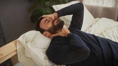 Yatak odasında sakallı, orta yaşlı bir adam yatakta uzanmış, boynunu tutarken rahatsızlık ya da acı içinde beliriyor..