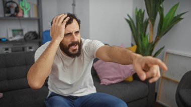 A confused man with a beard points and scratches his head in an indoor living room setting. clipart