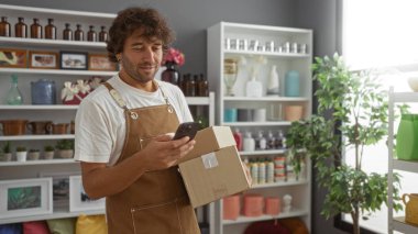Önlük giyen genç adam bir kutu tutuyor ve arka planda vazolar, şişeler ve bitkilerle dolu bir dekor dükkanında telefonunu kontrol ediyor.