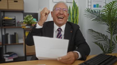 Modern bir ofiste başarıyı kutlayan, elinde bir belge tutan ve zaferle yumruğunu sallayan, takım elbiseli, yaşlı, beyaz bir adam..