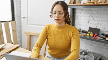 Marangozluk atölyesinde çalışan odaklanmış genç bir kadın, çeşitli aletler ve ahşap parçalarıyla çevrili..