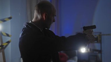 Elinde silah olan kel bir adam, kapalı alandaki karanlık bir suç mahallini dikkatle araştırıyor..