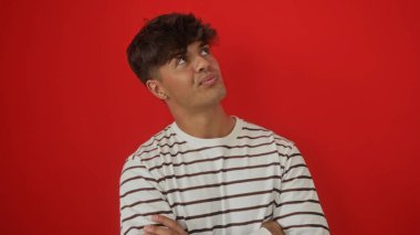 Çizgili tişörtlü yakışıklı bir İspanyol genç kollarını kavuşturur ve canlı kırmızı bir arka plana doğru bakar, içten ve düşünceli bir ifade yayar..