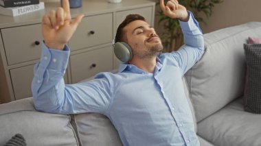 Sakallı, rahat bir oturma odasında dinlenen, kulaklıklı, evde rahat bir koltukta müzik dinleyen genç İspanyol bir adam..