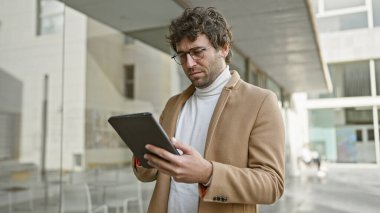 Odaklanmış sakallı bir İspanyol, şehir sokaklarında bir tablet kullanıyor, kentsel profesyonelliği ve modern teknolojiyi somutlaştırıyor..