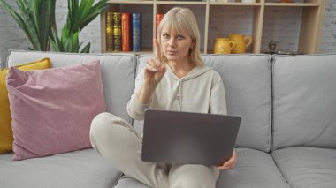 Modern bir oturma odasında dizüstü bilgisayarda çalışırken el kol hareketi yapan sarışın kadın..