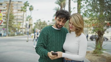 Gülümseyen bir çift palmiye ağaçlarıyla dolu kalabalık bir caddede akıllı bir telefonu paylaşırken kucaklaşır..